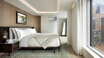 A modern hotel room with a large bed, a nightstand, a mirror, and a view of the city through large windows.