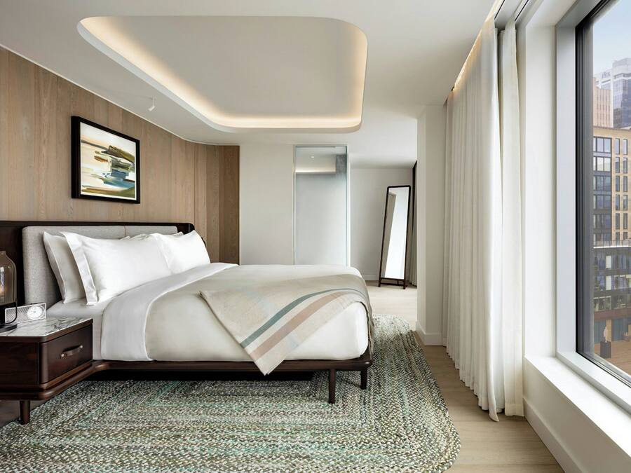 A modern hotel room with a large bed, a nightstand, a mirror, and a view of the city through large windows.