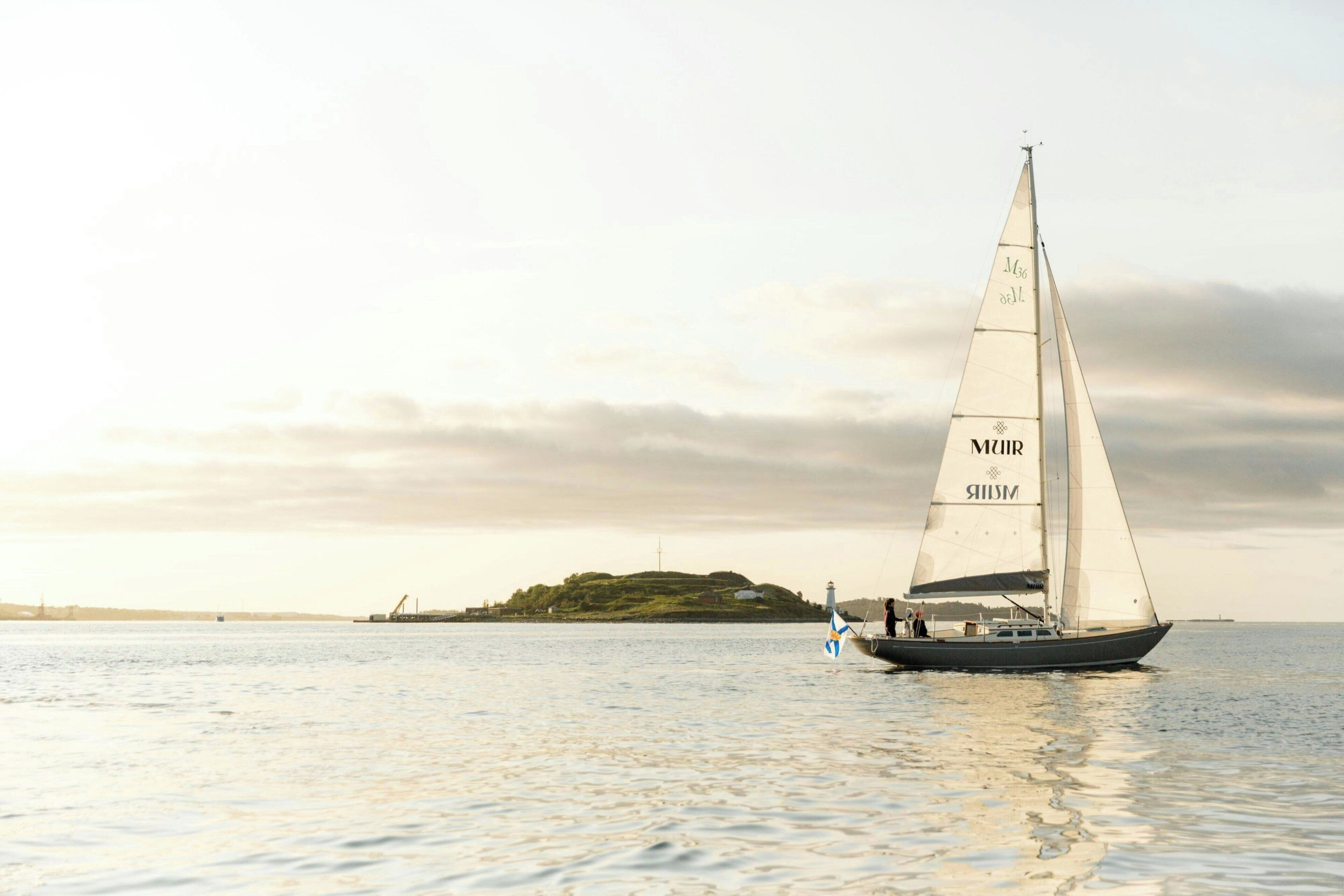 A sailboat with "MUR" and "MUS" on its sails, sailing on calm waters with a distant island and lighthouse in the background.