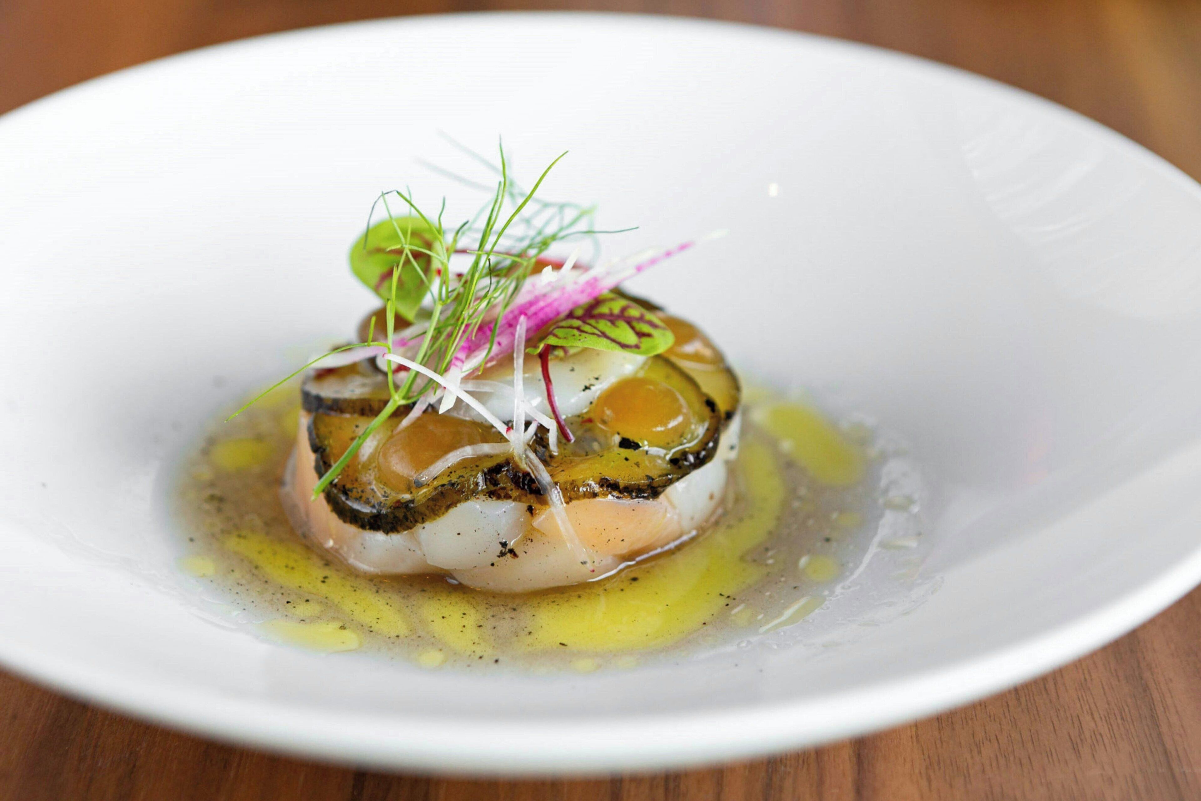 Un plato blanco con un guiso de mariscos, adornado con vegetales verdes y una salsa amarilla.
