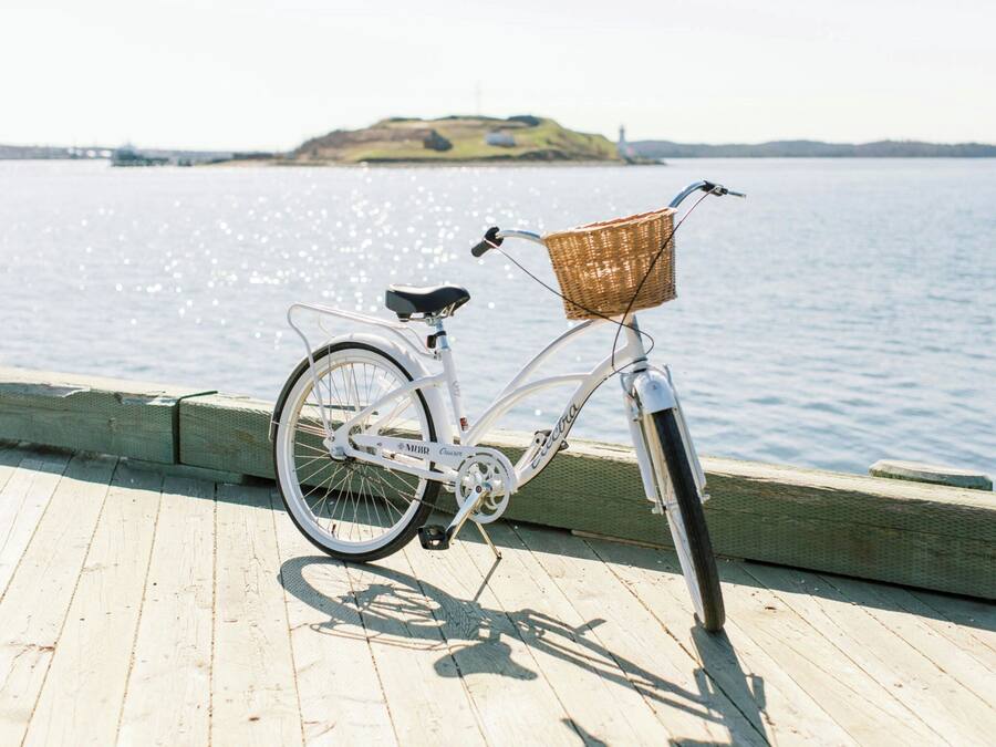 A white bicycle with a wicker basket on a wooden dock by a calm body of water.