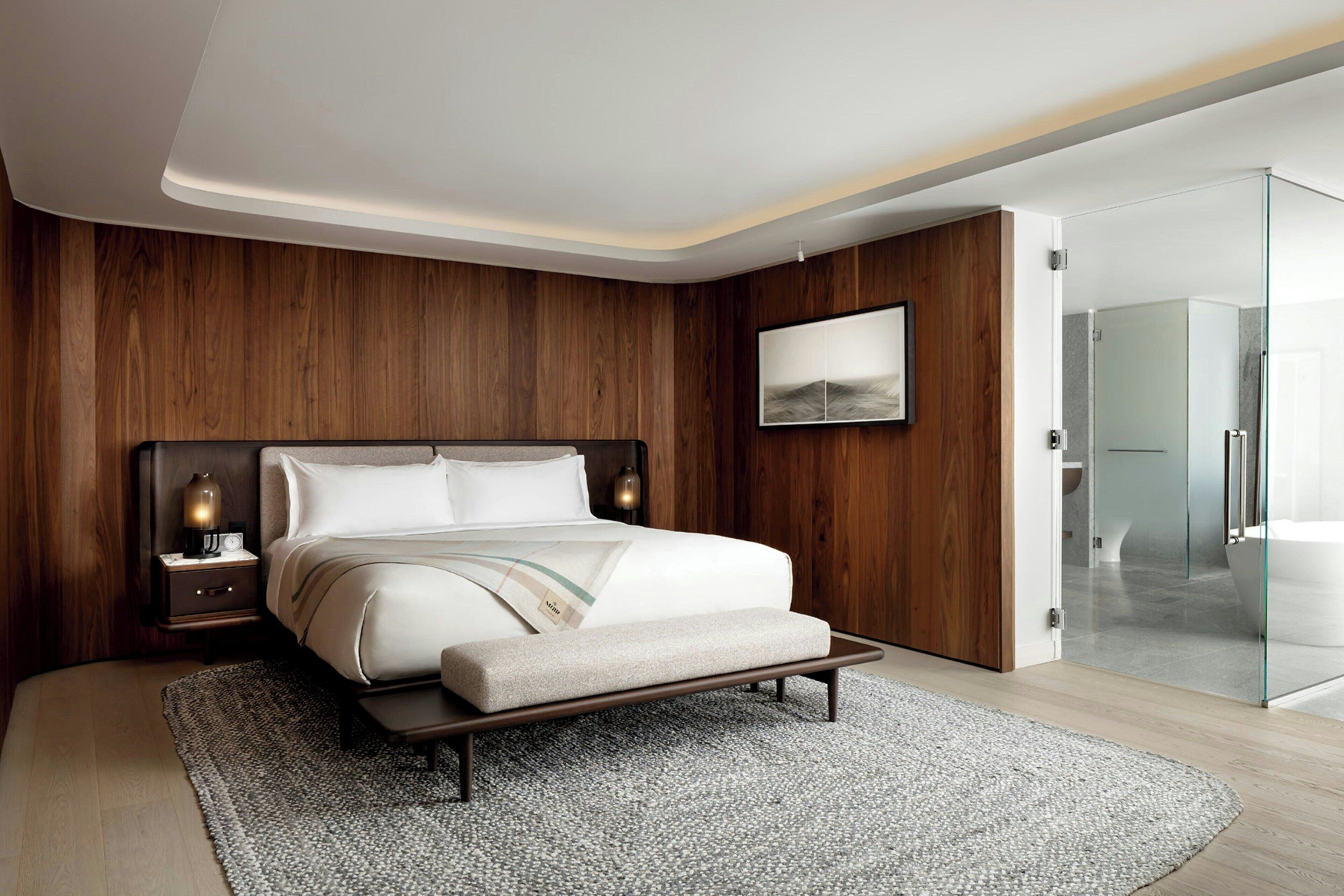 A modern bedroom with a wooden headboard, a bedside table with a lamp, a large bed with a bench, and a glass shower area.