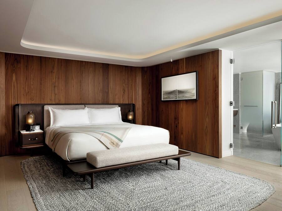 Un dormitorio moderno con cabecera de madera, una mesita de noche con lámpara, una cama grande con banco y una zona de ducha de vidrio.