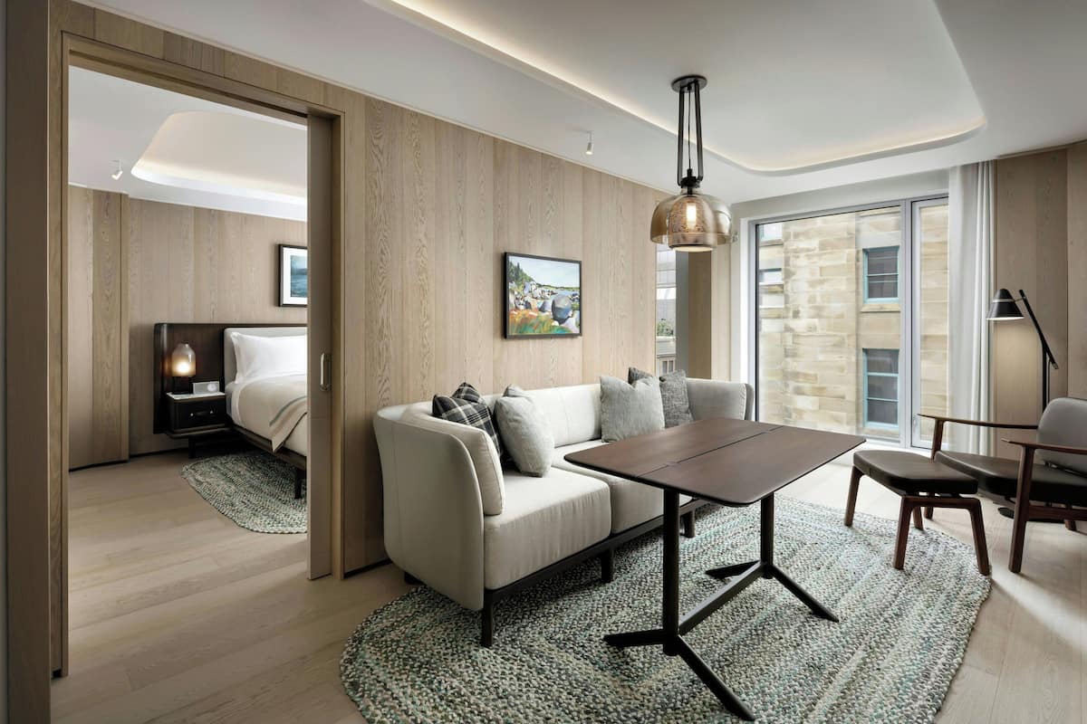 A modern living room with a sofa, a coffee table, and a dining area. There is a bed in the background, a large window, and a door leading to another room.