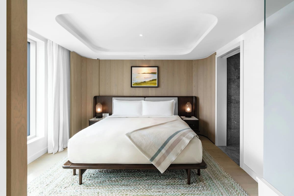 A modern hotel room with a large bed, wooden panel walls, a painting above the bed, and a nightstand with a lamp.