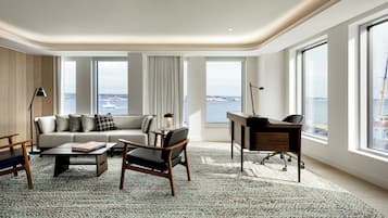 Uma sala de estar moderna com sofá, mesa de centro e poltrona, grandes janelas com vista para o mar e uma escrivaninha com cadeira.