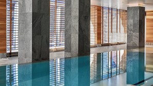 Un área de piscina interior moderna con suelo de vidrio transparente, grandes columnas de piedra y paredes de listones de madera que dejan pasar la luz natural.