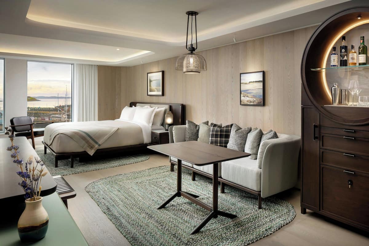 A modern hotel room with a bed, sofa, dining table, and a bar area.