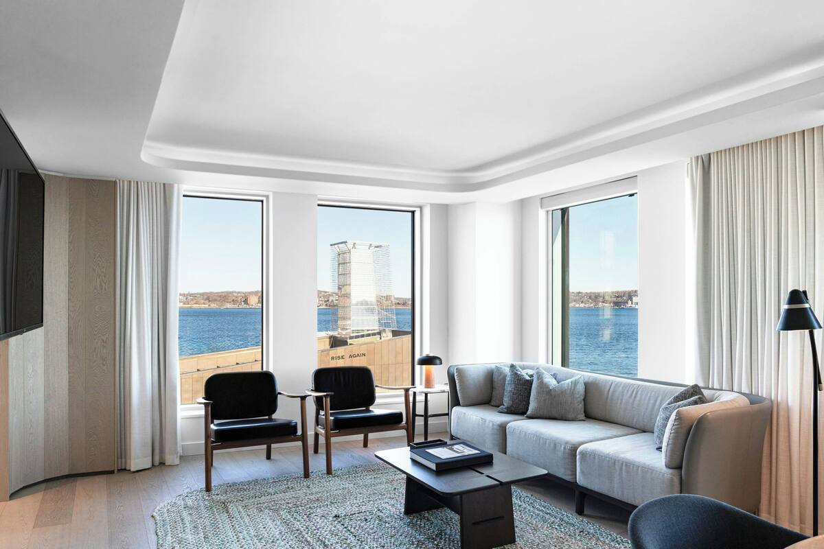A modern hotel room with a large window offering a view of the sea and a distant cityscape. There is a sofa, a chair, and a small table with a lamp.