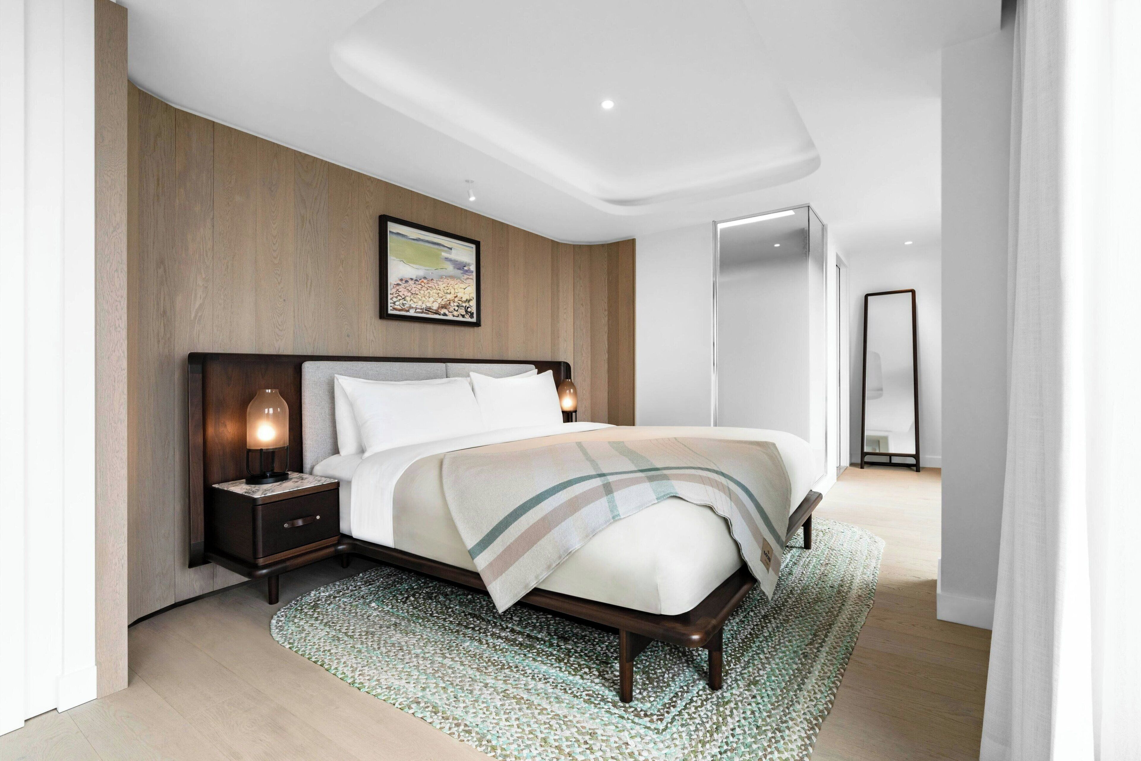 A modern hotel bedroom with a wooden headboard, a large bed with a plaid blanket, a bedside table with a lamp, and a framed picture on the wall.