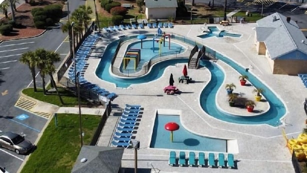 Vista aérea de un complejo de piscinas con múltiples carriles, sillas de descanso y una zona de juegos pequeña.