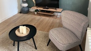 Ein modernes Wohnzimmer mit einem Flachbildfernseher, einem runden schwarzen Couchtisch mit Kerzen, einem gemusterten Sessel, einem Holzregal mit Küchenutensilien und einer weißen Stehlampe.