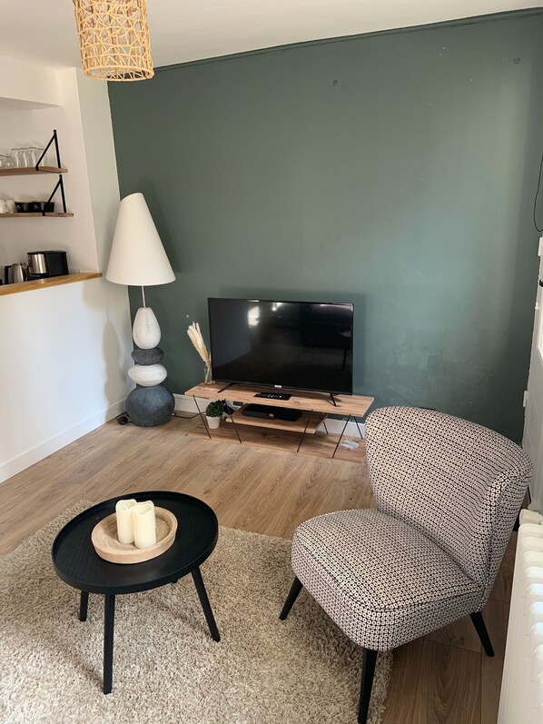 Ett modernt vardagsrum med en platt-TV, ett runt svart soffbord med ljus, en mönstrad fåtölj, en hylla i trä med köksartiklar och en vit golvlampa.