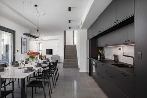 Eine moderne Küche mit dunklen Schränken, ein Essbereich mit einem langen Tisch und Stühlen sowie eine Treppe zu einer oberen Ebene.