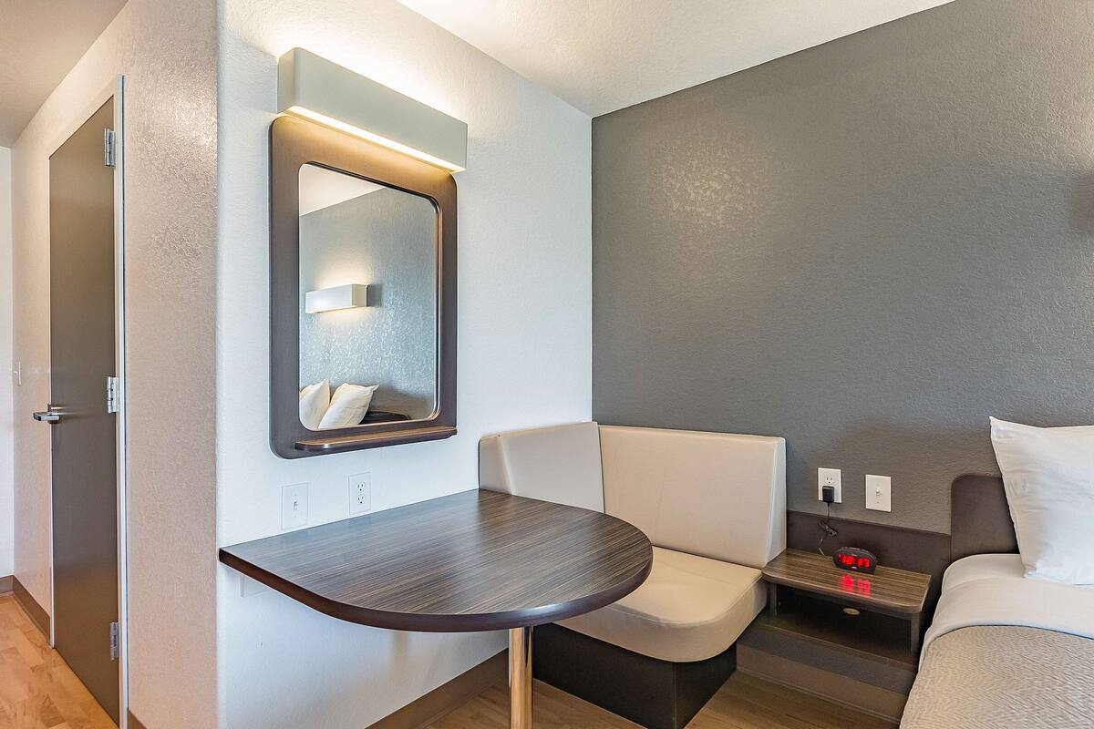 A compact hotel room with a small table, a cushioned bench, a bed, a nightstand with a lamp, and a mirror.