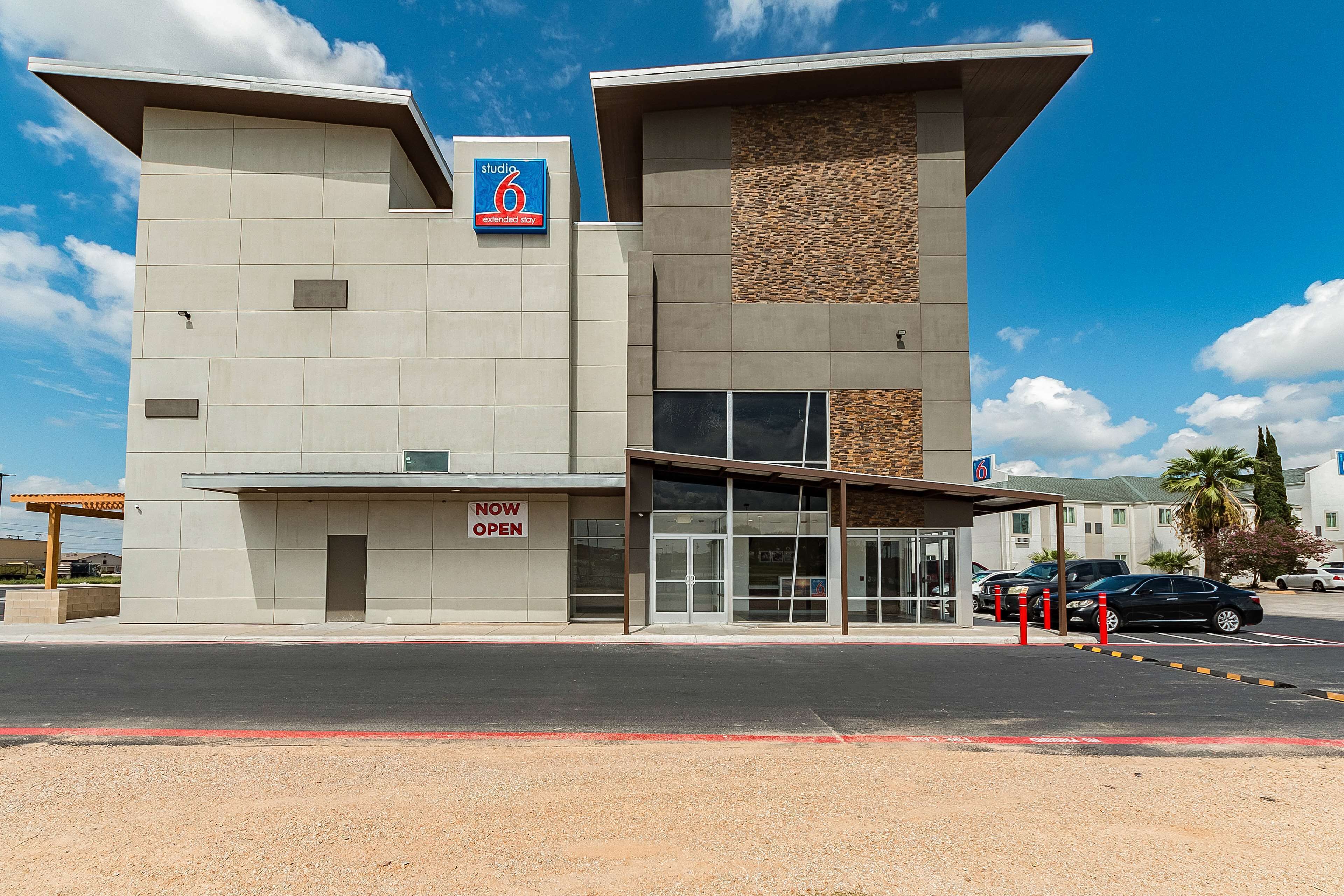 A modern building with a 'Studio 6' sign, a 'Now Open' sign, and a parking area with cars.