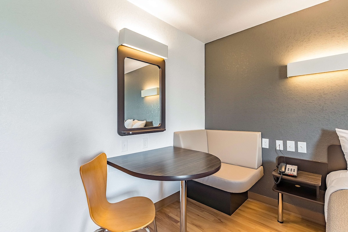 A modern hotel room with a round table, a chair, a sofa, a bedside table with a phone, and a mirror.