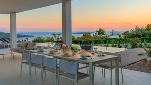 Een eetruimte met een tafel voor vier personen, met uitzicht op een schilderachtig zee- en heuvellandschap tijdens de zonsondergang.