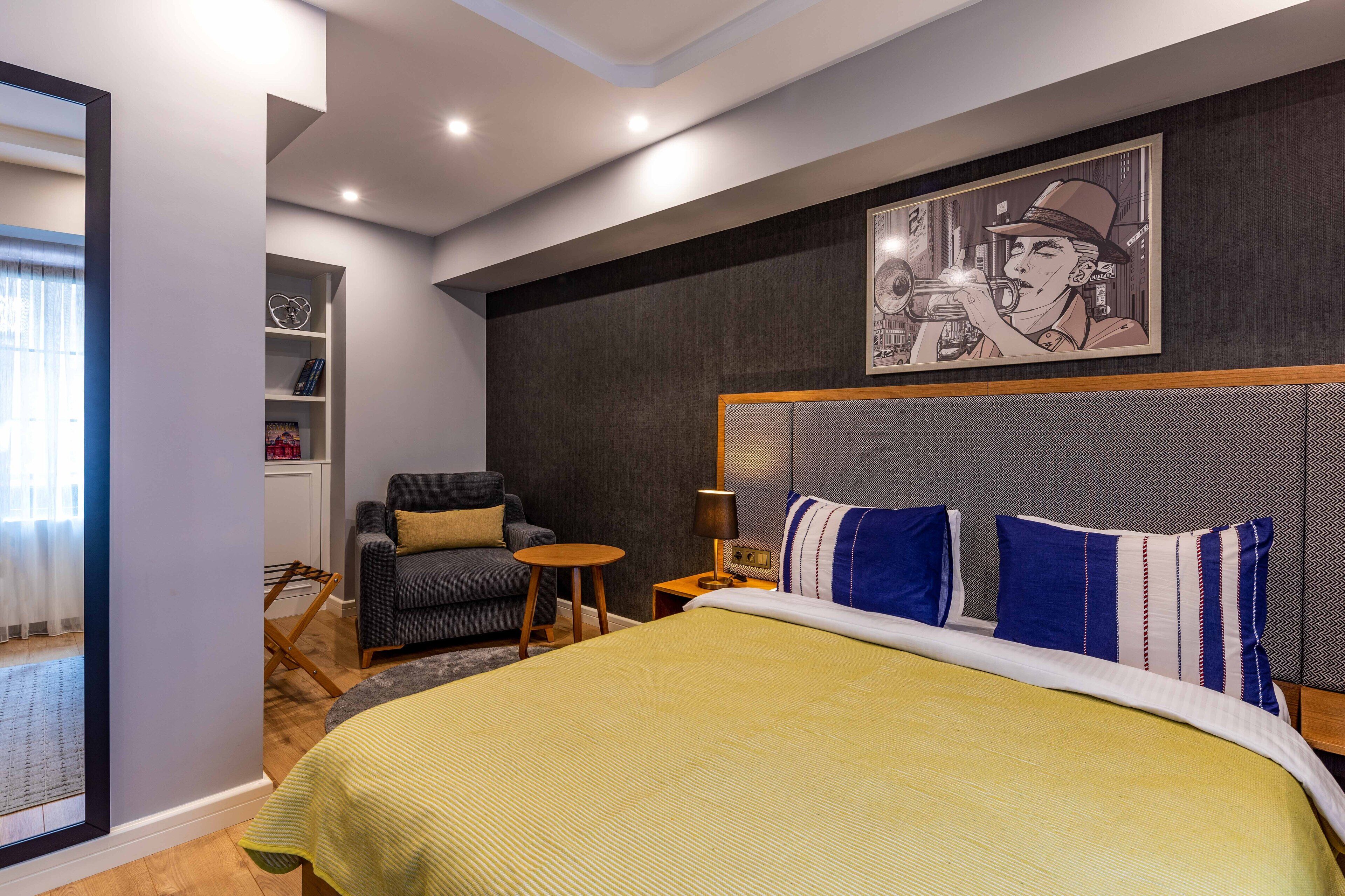 Ein modernes Hotelzimmer mit einem großen Bett, einem grauen Sessel, einem kleinen runden Tisch und einem an der Wand hängenden Kunstwerk.