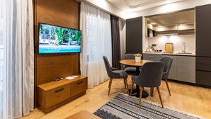 Ein modernes Hotelzimmer mit einem an einer Holzvertäfelung montierten Fernseher, einem Essbereich mit rundem Tisch und Stühlen sowie einer Küchenzeile mit Spüle und Schränken.