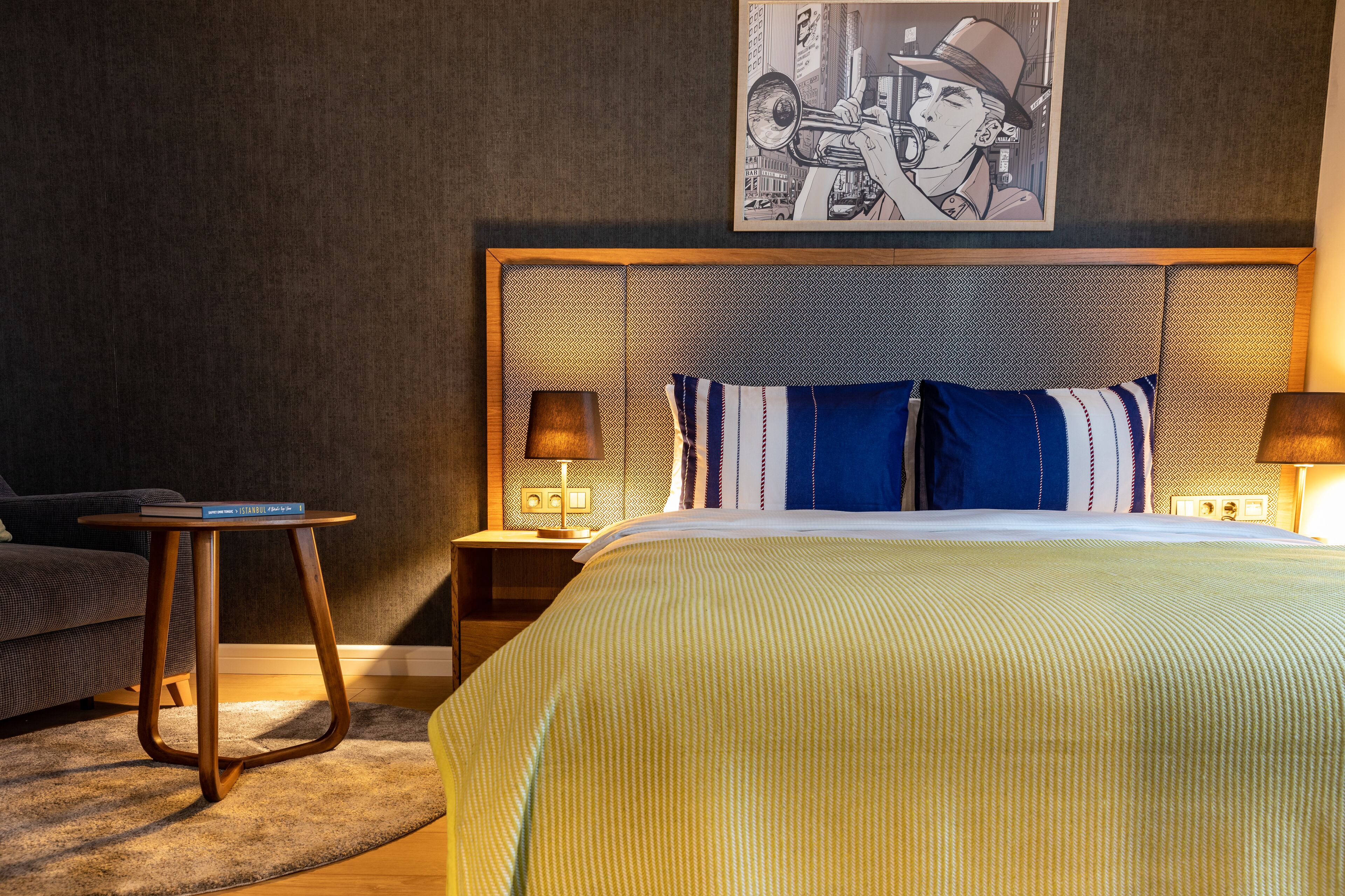 Ein Hotelzimmer mit einem großen Bett, einem Nachttisch, einer Lampe, einem gerahmten Bild an der Wand und einem kleinen Beistelltisch.