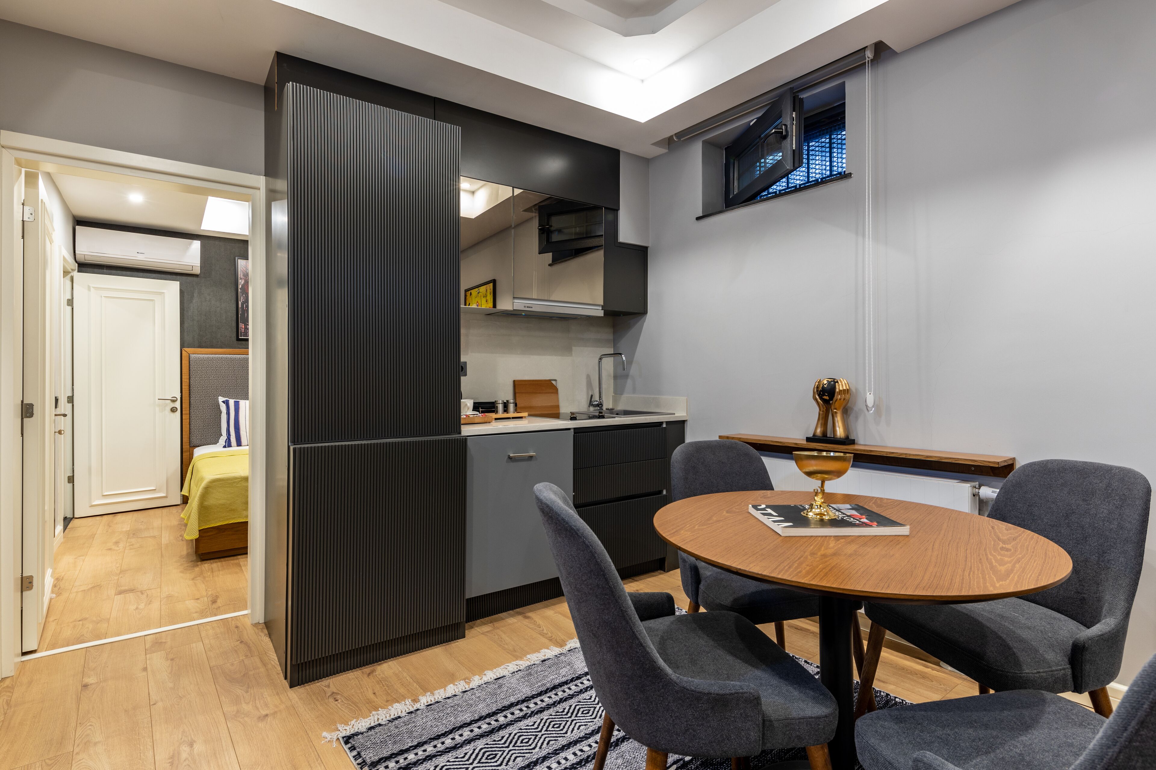 Een moderne keuken met grijze kastjes, een houten eettafel en een klein raam.