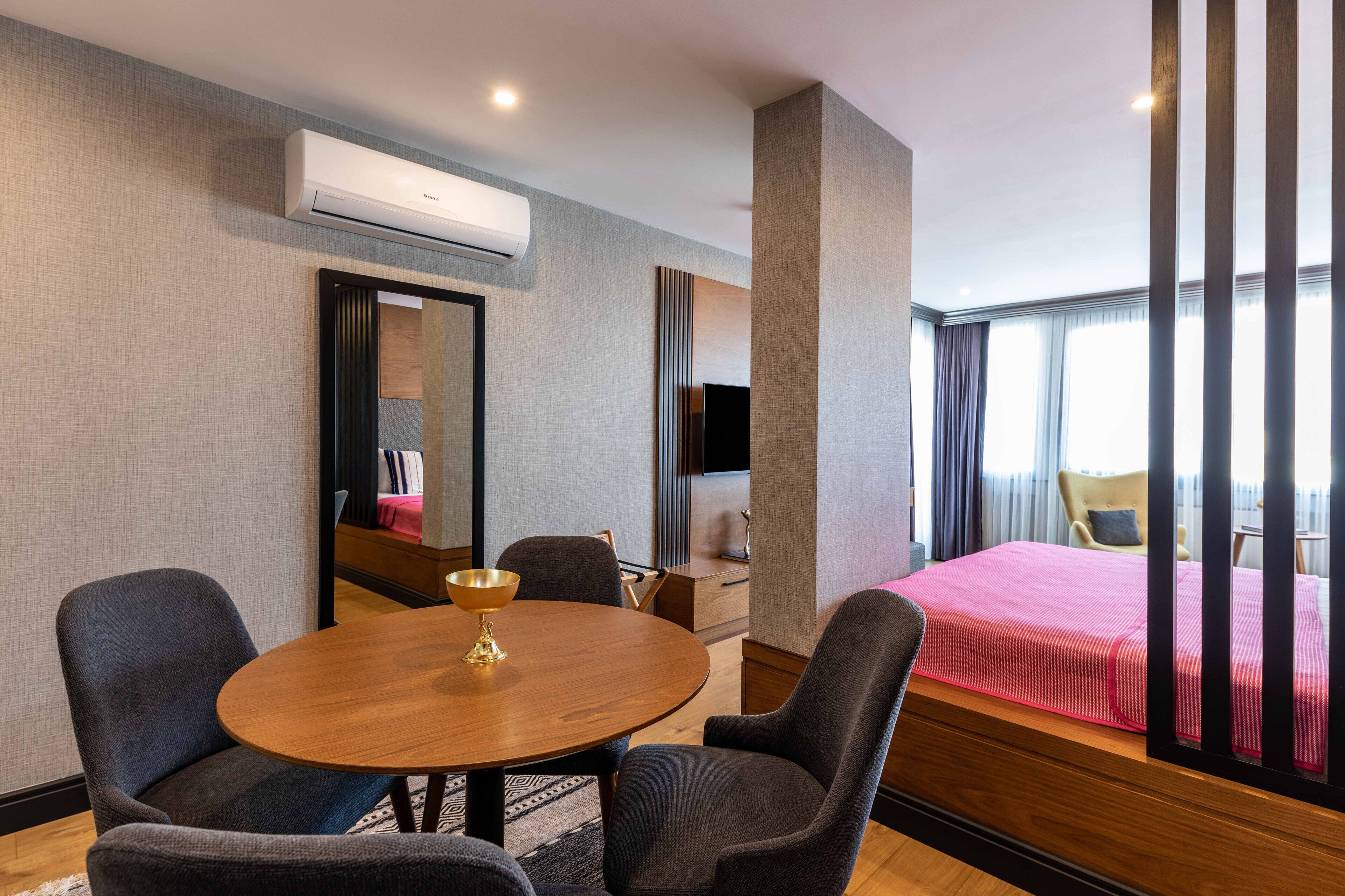 A hotel room with a round wooden table, two armchairs, a bed with a red bedspread, a TV, and an air conditioning unit.
