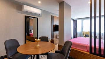 A hotel room with a round wooden table, two armchairs, a bed with a red bedspread, a TV, and an air conditioning unit.
