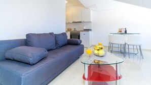 Un salon moderne avec un canapé gris, une table basse en verre avec un pied rouge, et un coin repas avec des chaises blanches et une table en verre.