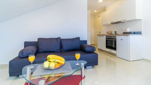 Un salon moderne avec un canapé gris, une table basse en verre sur laquelle sont disposés des fruits et des boissons, et une cuisine en arrière-plan.