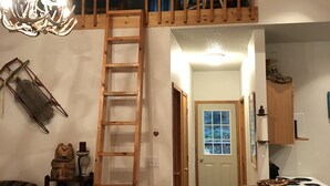 A loft bedroom with a wooden ladder leading to a lofted sleeping area, a chandelier, and a kitchen area with a bar stool. - Rapid Creek Get Away (Hill City)