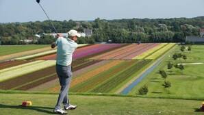 Ein Golfplatz mit einem Spieler im Schwung, dahinter farbenfrohe Blumenfelder und ein klarer Himmel.
