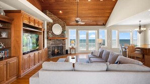 Ein geräumiges Wohnzimmer mit großem Fernseher, Holzsärchen, einem Steinkamin und Meerblick durch große Fenster.