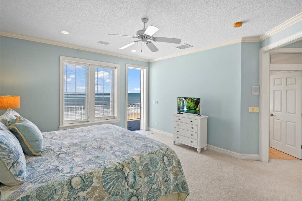 A bedroom with a bed, a TV, a ceiling fan, and a view of the ocean through large windows.