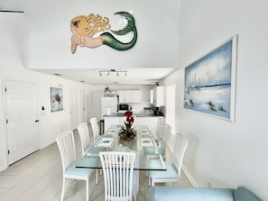 Uma sala de jantar moderna com mesa de vidro, cadeiras brancas e uma cozinha ao fundo.