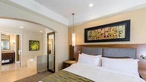 Une chambre d’hôtel avec un grand lit, une tête de lit, une table de chevet, une petite table, une œuvre d’art accrochée au mur et une porte donnant accès à la salle de bain.