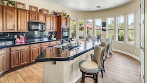 A modern kitchen with wooden cabinets, a black countertop, and a large island. There are bar stools and a dining area with a view of the outdoors. - Oasis in the desert - your hiking and biking paradise. (Scottsdale)