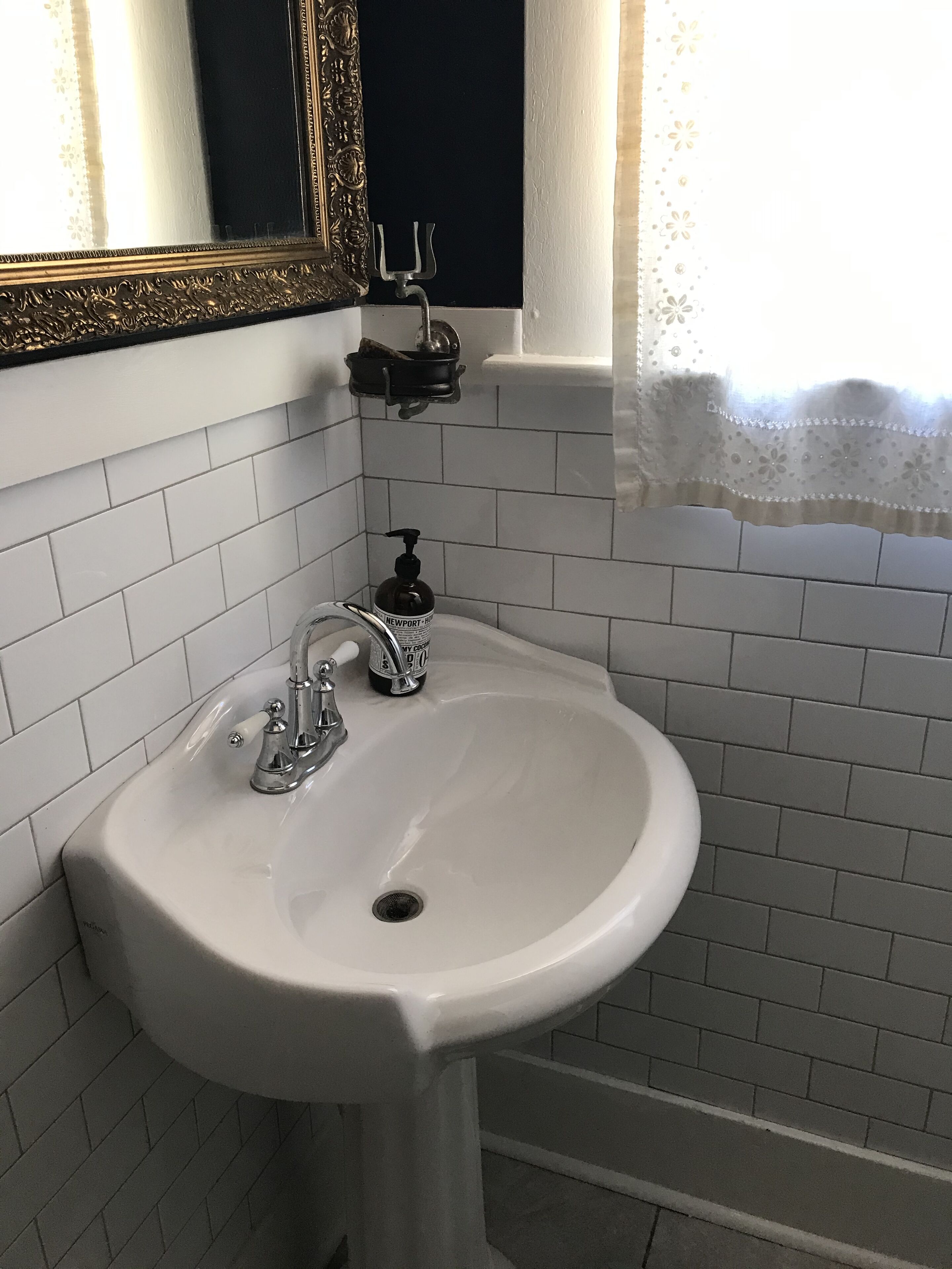 Un baño con un lavamanos blanco, un espejo con un marco ornamentado y una ventana con cortina de encaje.