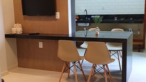 A modern kitchen with a flat-screen TV mounted on a wooden panel, a black countertop, and a white tiled backsplash. There is a dining area with two chairs and a glass-top table.