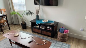Ein Wohnzimmer mit Couchtisch aus Holz, Wandfernseher, Bücherregal, Blumentopf und Korbwannenstuhl.