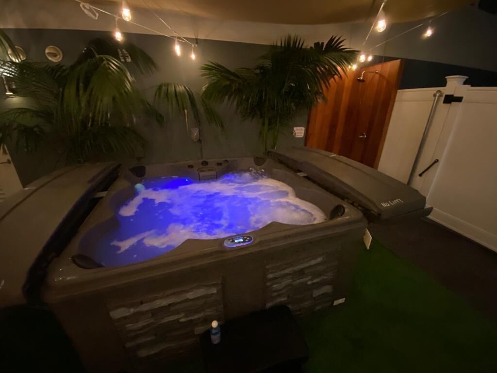 Uma banheira de hidromassagem iluminada por uma luz azul, cercada por plantas de palmeira, em um quarto com porta de madeira e parede branca.