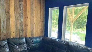 A corner room with a black leather sectional sofa, wooden panel wall, and large windows with a view of greenery. - Barn and bling on Windrock (Oliver Springs)
