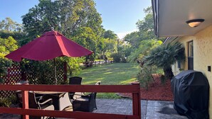 Eine Terrasse mit Gartenmöbeln, einem überdachten Grill und Blick auf einen Rasen mit Bäumen.