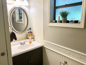 Une salle de bain avec un lavabo blanc, des meubles sombres, un miroir et une fenêtre agrémentée d’une plante et d’objets décoratifs.