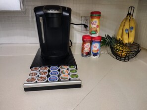 Une machine à café, plusieurs dosettes de café, une boîte de café au goût de noisette, une grappe de bananes et un ananas sur un plan de travail de cuisine.