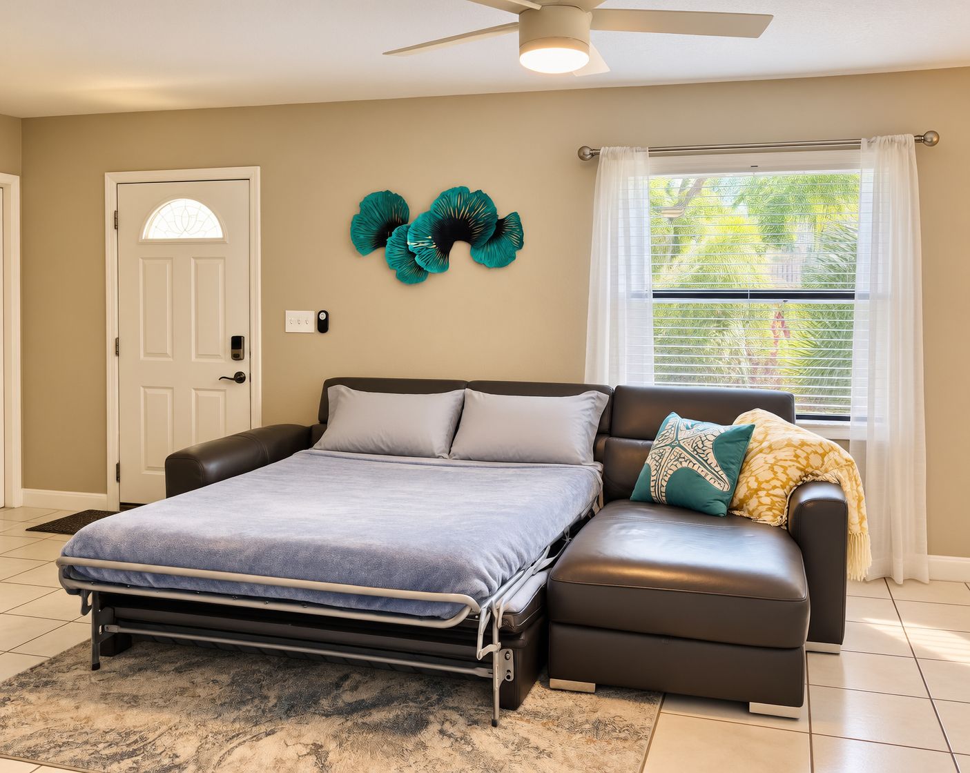 A bedroom with a bed, a sofa, a ceiling fan, a window with blinds, and a door.