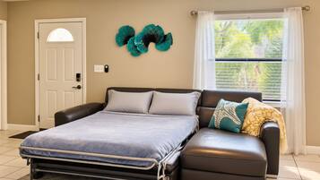 A bedroom with a bed, a sofa, a ceiling fan, a window with blinds, and a door.