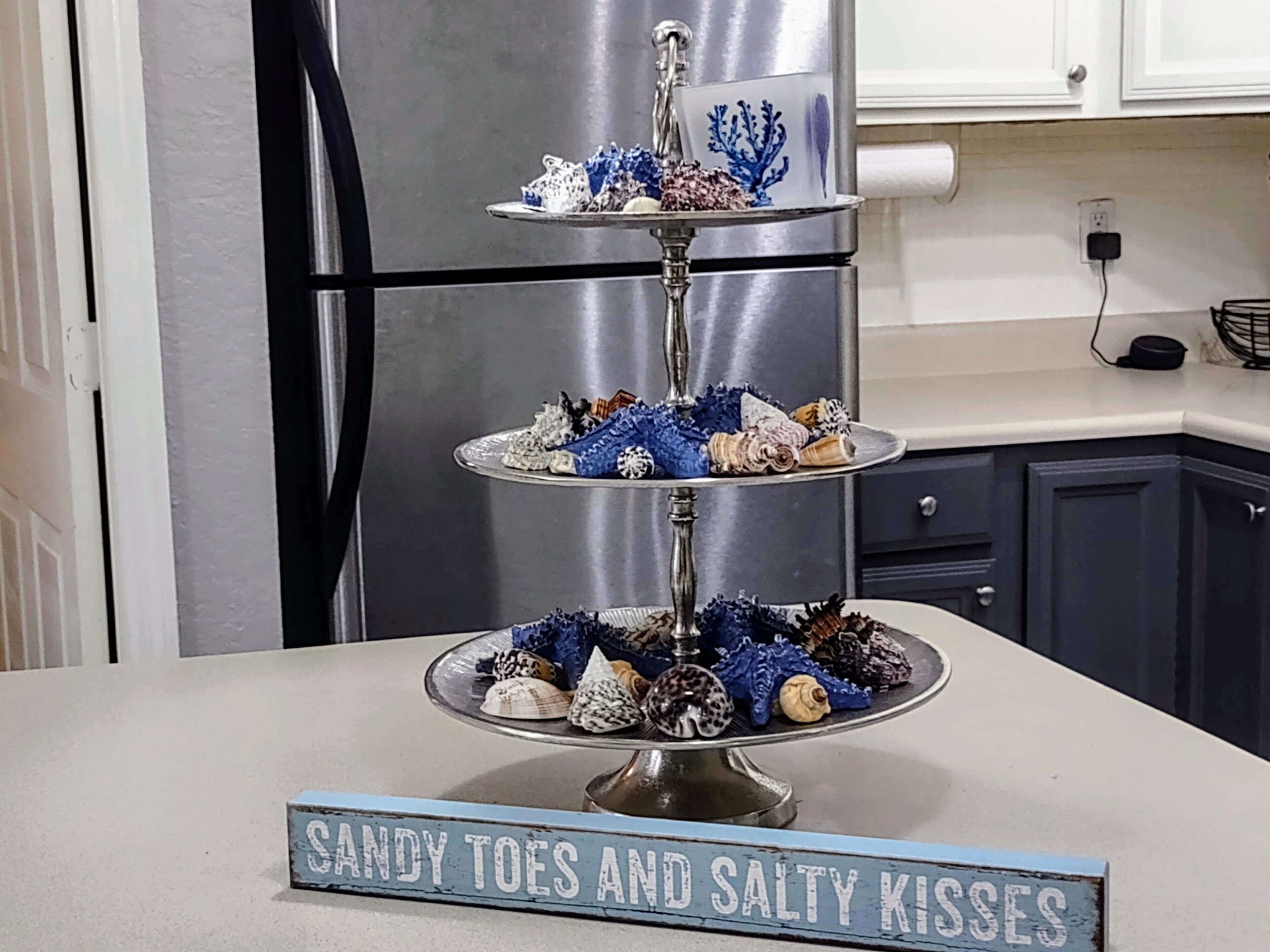 Un présentoir à trois niveaux pour desserts, avec une pancarte indiquant « SABLE SOUS LES PIEDS ET B AISERS SALÉS », dans une cuisine.