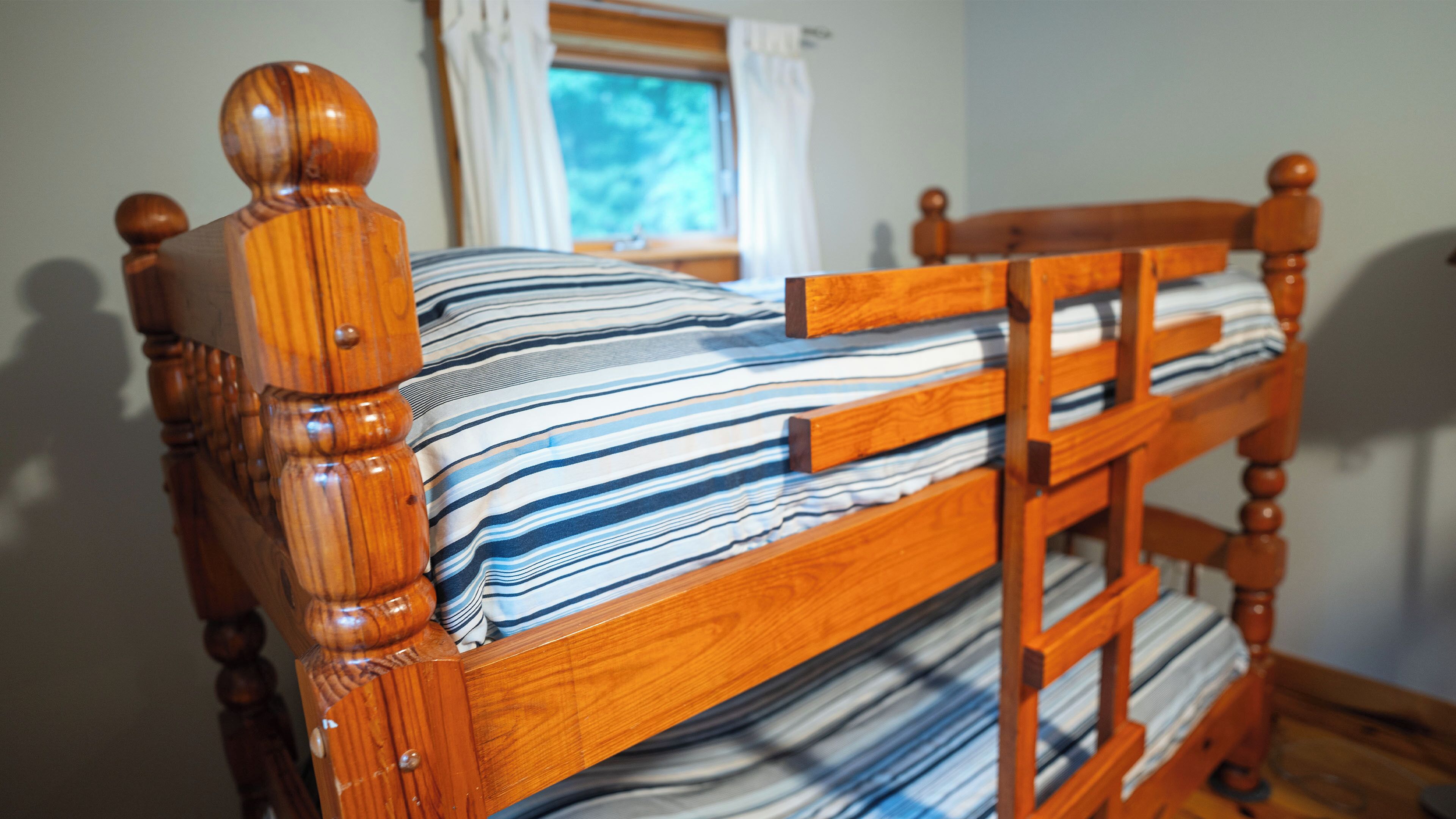 Un lit superposé en bois avec un matelas à rayures, une échelle en bois et une fenêtre avec des rideaux.
