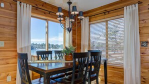 A wooden cabin dining room with a table set for four, large windows, and a chandelier. - Maple Cove Lodge on Lake Sebago Waterfront! (Casco)